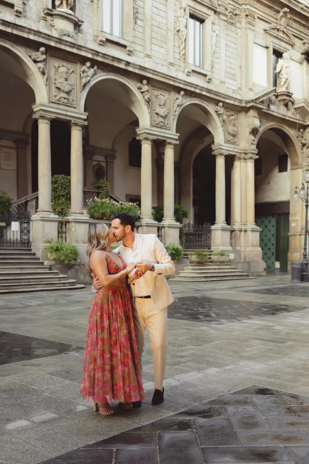 On the wet stone paving of Piazza dei Mercanti, the couple kisses in a close embrace before a colonnaded Renaissance arcade.