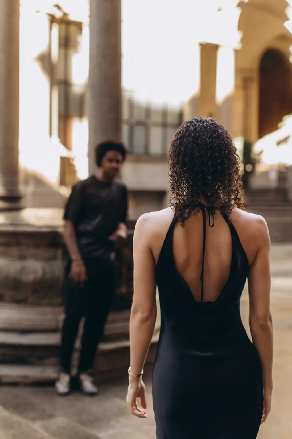 Seen from behind, a woman in an open-back black dress walks toward her partner waiting by the steps in Piazza dei Mercanti.