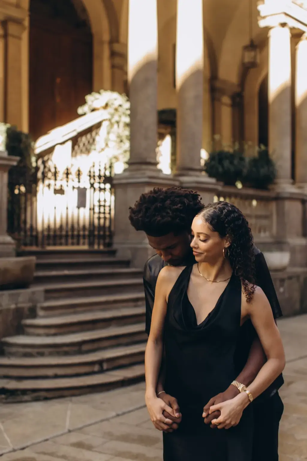 A man hugs his partner from behind, his face hidden in her curls, at the foot of the stone staircase in Piazza dei Mercanti.