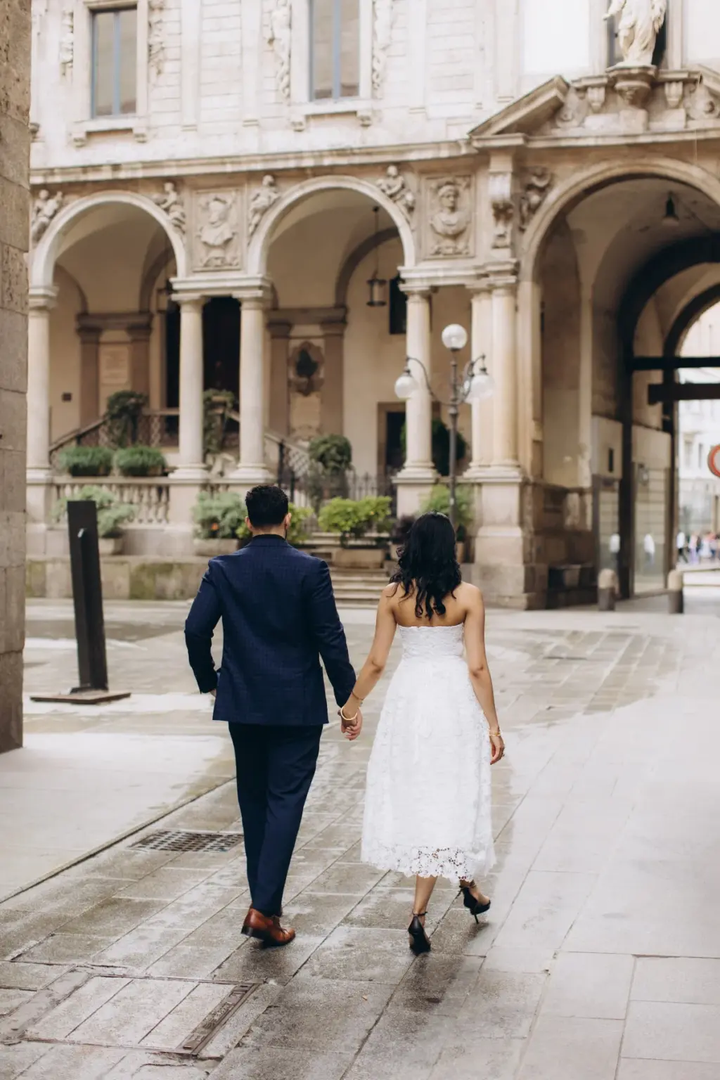 From behind, the couple strolls hand in hand toward the arcaded palazzo of Piazza dei Mercanti, her white lace hem swaying.