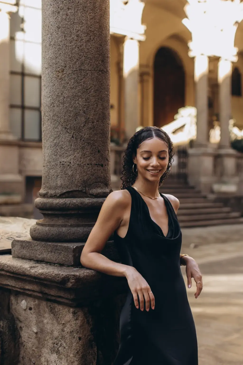 A woman in a black halter dress leans on a granite column under the arcades of Piazza dei Mercanti, smiling down at the ground.