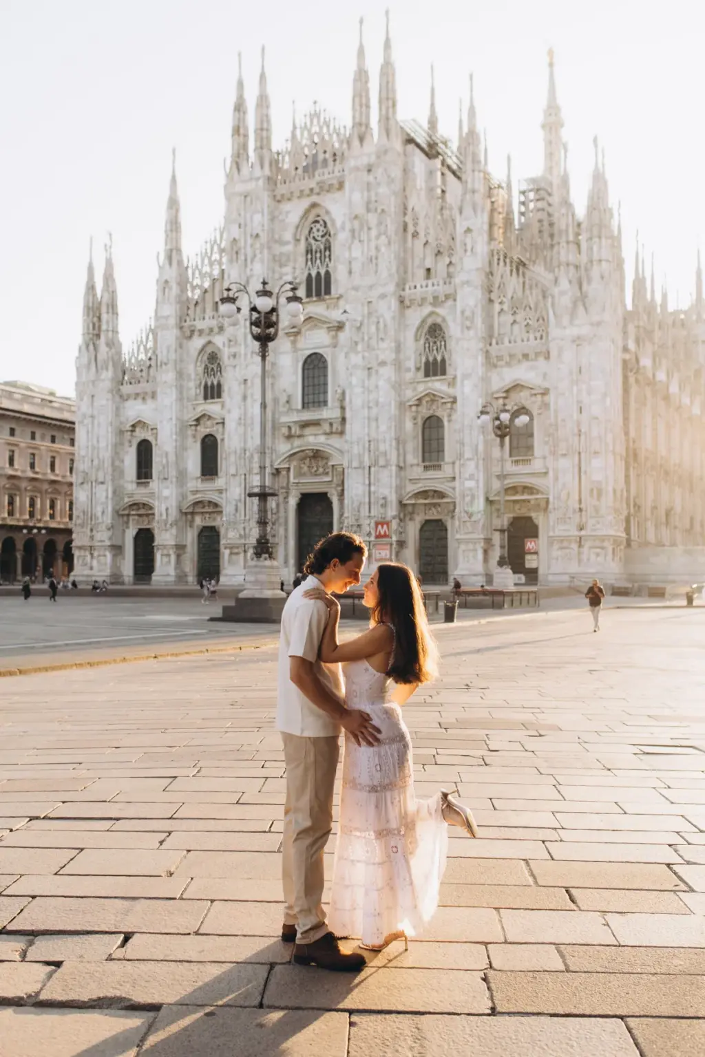 A woman in a white lace dress lifts one foot as she kisses her partner in the open Piazza del Duomo before the cathedral.