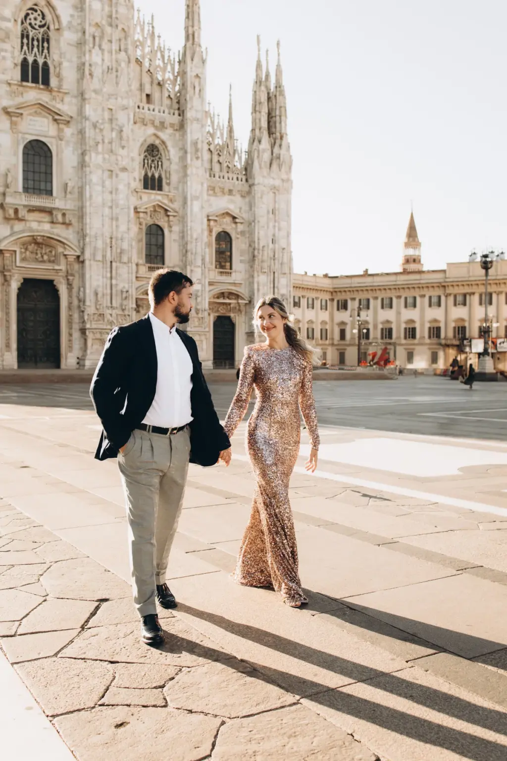 Hand in hand, a man in a blazer and a woman in a long sequin gown walk across the sunlit Piazza del Duomo, the cathedral at left.