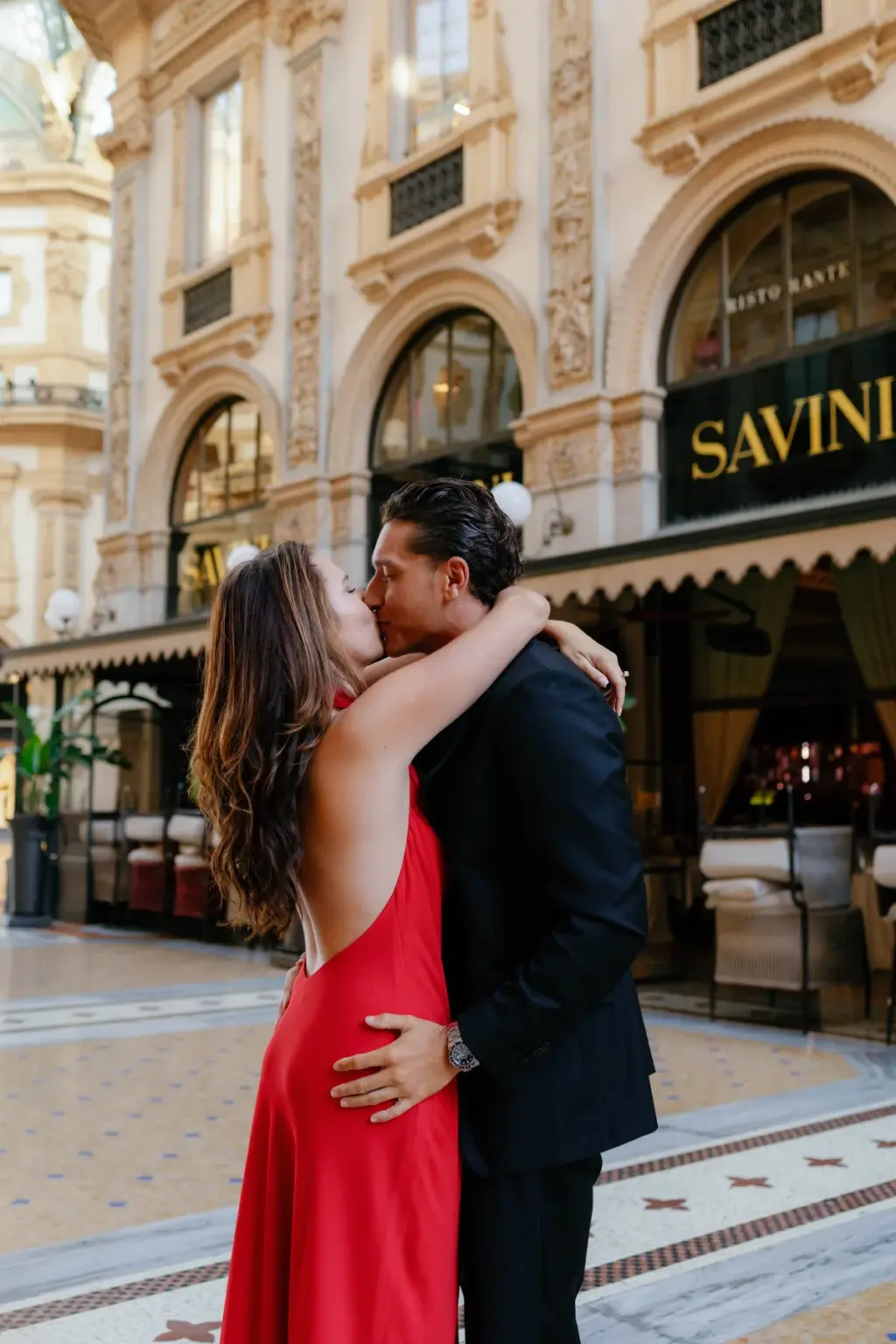 Arms wrapped around his neck, she kisses him outside Savini under the gilded shop arches of Galleria Vittorio Emanuele II.