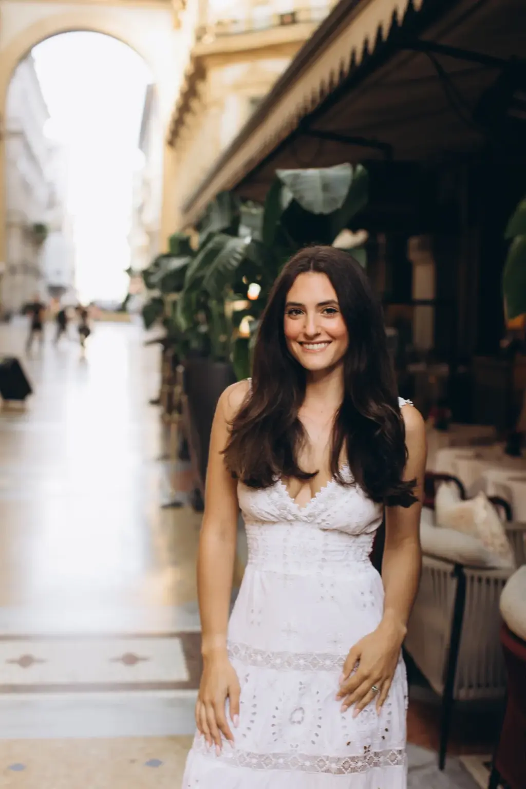 A smiling woman with long dark hair stands in a white lace dress beside the Savini terrace and its tall palms inside the Galleria.