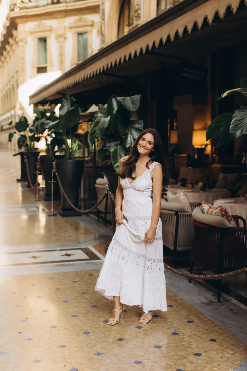 Full-length, a woman in a white tiered dress and sandals smiles on the mosaic floor of the Galleria by the Savini awning.