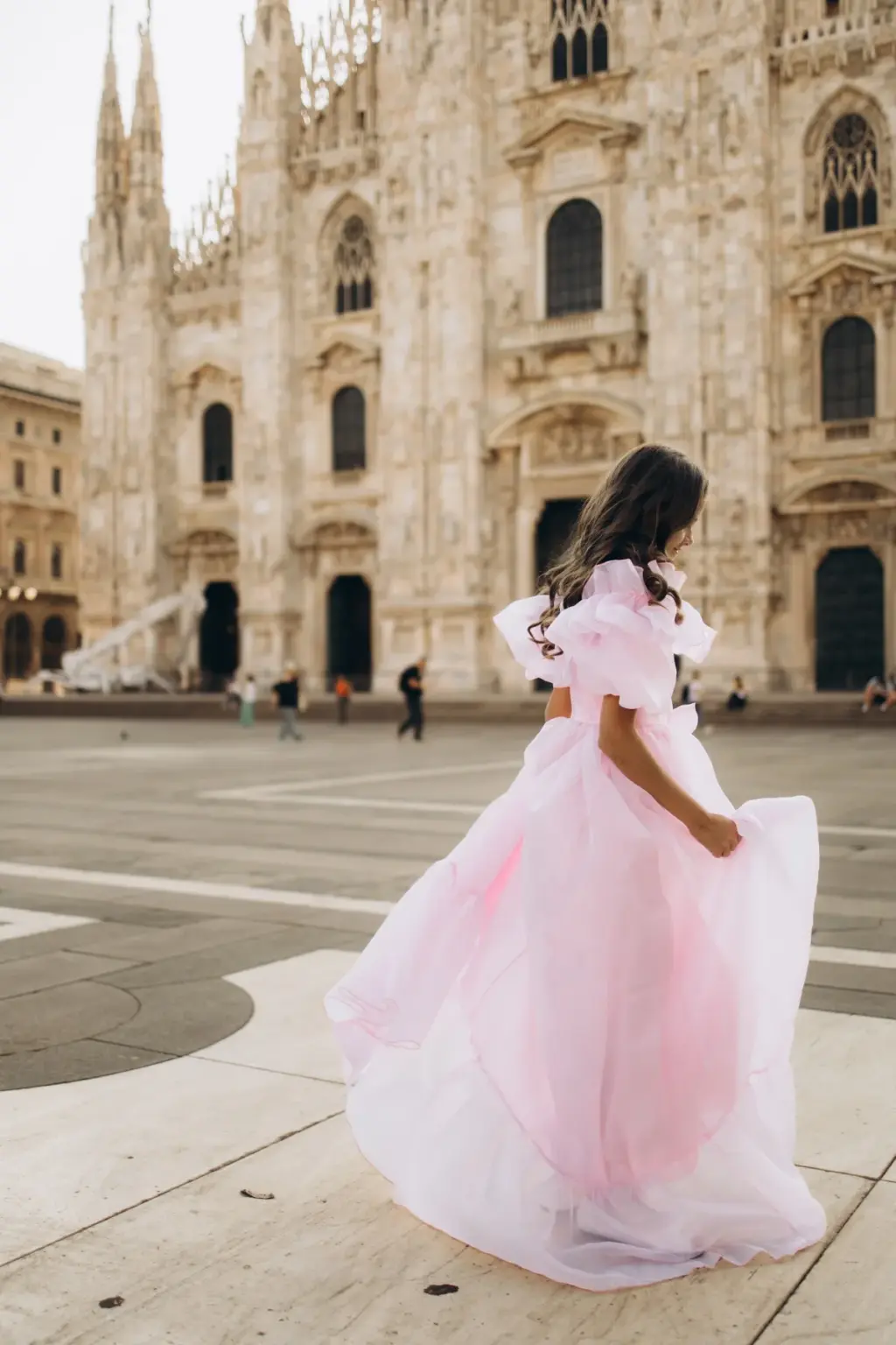 A girl in a long pink ruffled gown spins and looks back over her shoulder on the paved square in front of the Duomo.