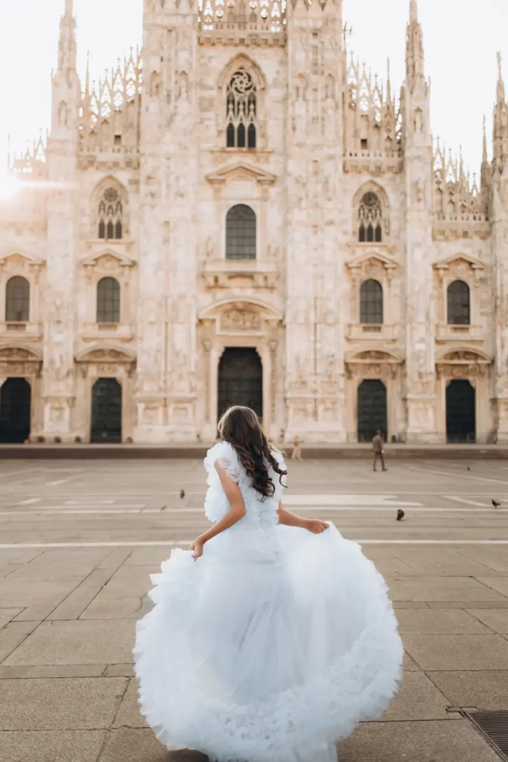 Seen from behind, a girl in a white tulle dress runs toward the Duomo di Milano across the open piazza, the ruffles flying.