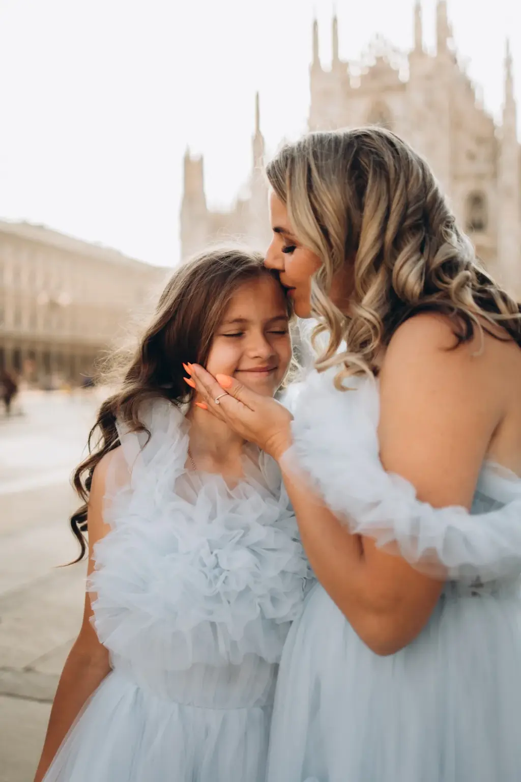 A mother kisses her smiling daughter's forehead, both in pale blue tulle, with the Duomo spires softly blurred behind them.