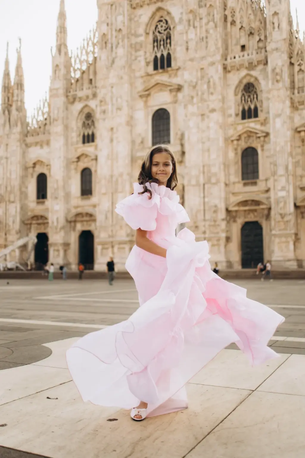 A smiling girl in a pink ruffled gown swirls her skirt wide on Piazza del Duomo, the white cathedral facade filling the frame.