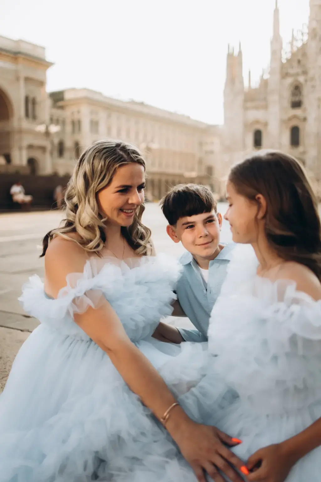 A mother sits close with her son and daughter on the Duomo square, all smiling at each other, the cathedral spires behind.