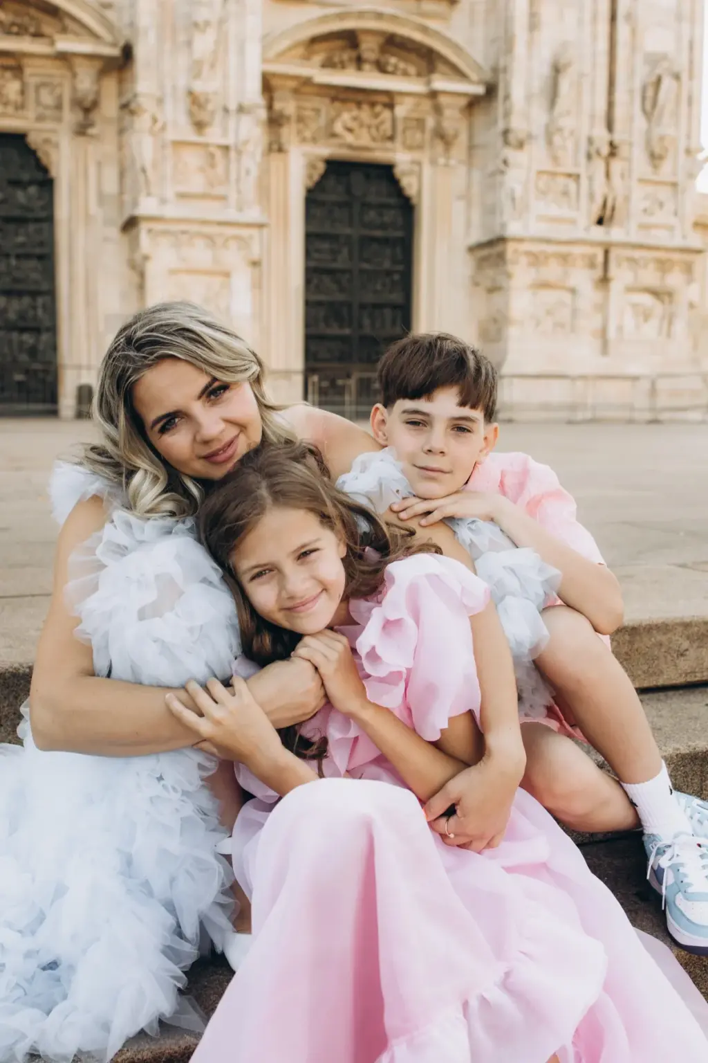 A mother sits on the Duomo steps with her arms around her daughter in pink and her son leaning on her shoulder.