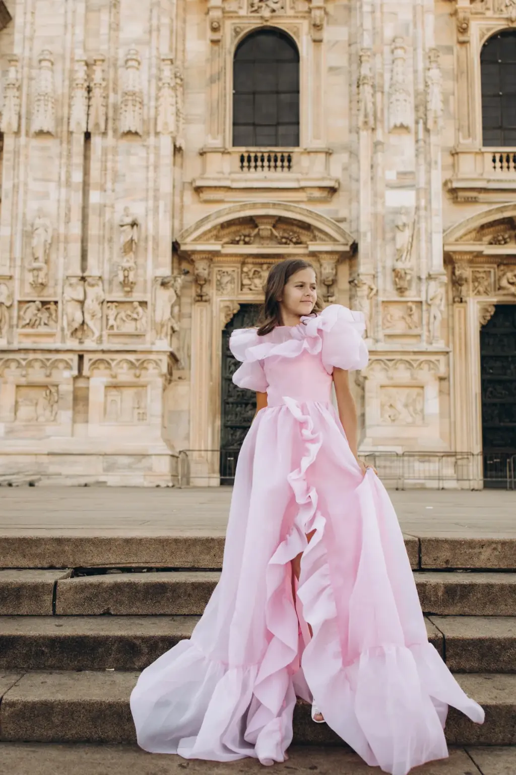 A girl in a pink ruffled gown stands on the stone steps of the Duomo di Milano below a carved doorway, the train spread on the stairs.