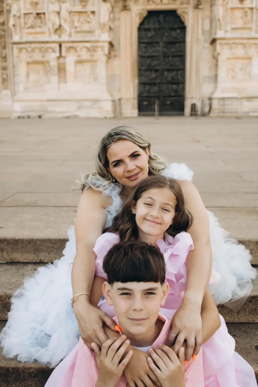 A mother, daughter and son stack up in a hug on the Duomo steps, the boy in front in a pink shirt, the bronze doors behind.
