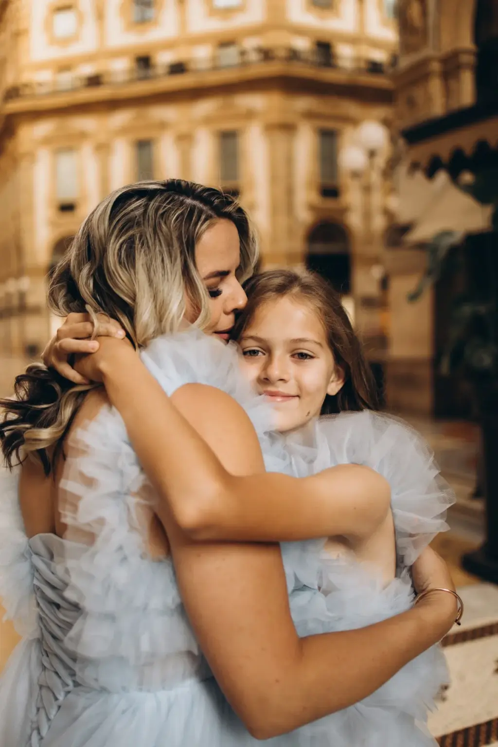 A daughter rests her head on her mother's shoulder in a tight hug, both in light blue tulle, with Galleria shopfronts blurred behind.