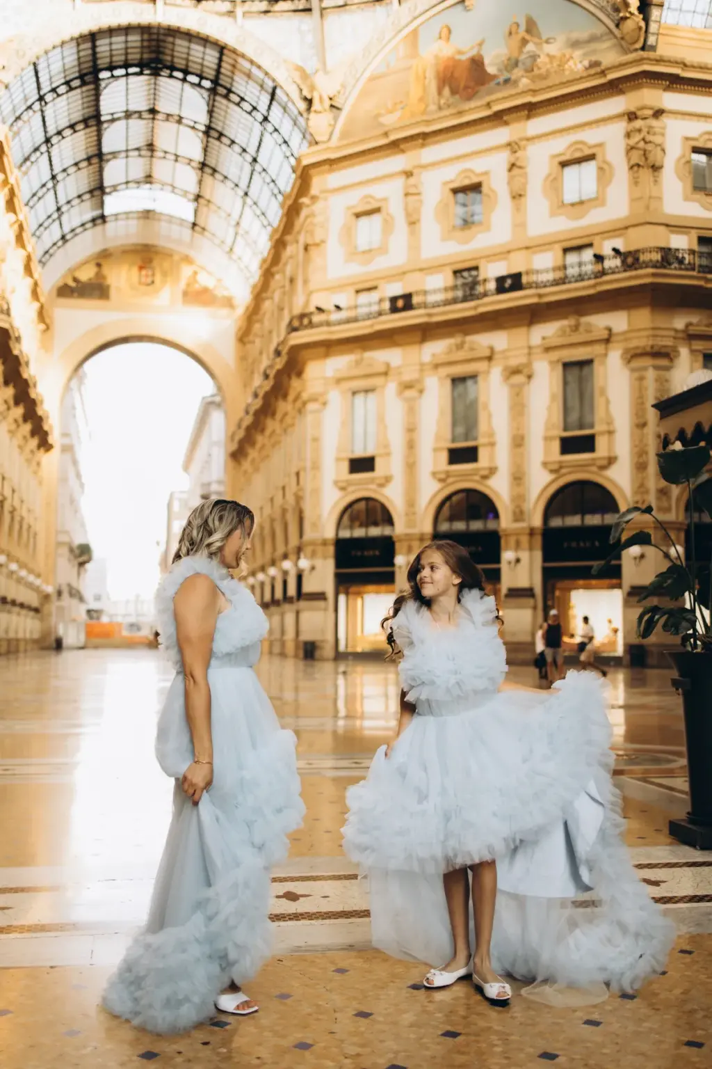 A mother and her daughter in ruffled blue tulle gowns stand facing each other under the Galleria's glass dome and painted lunettes.