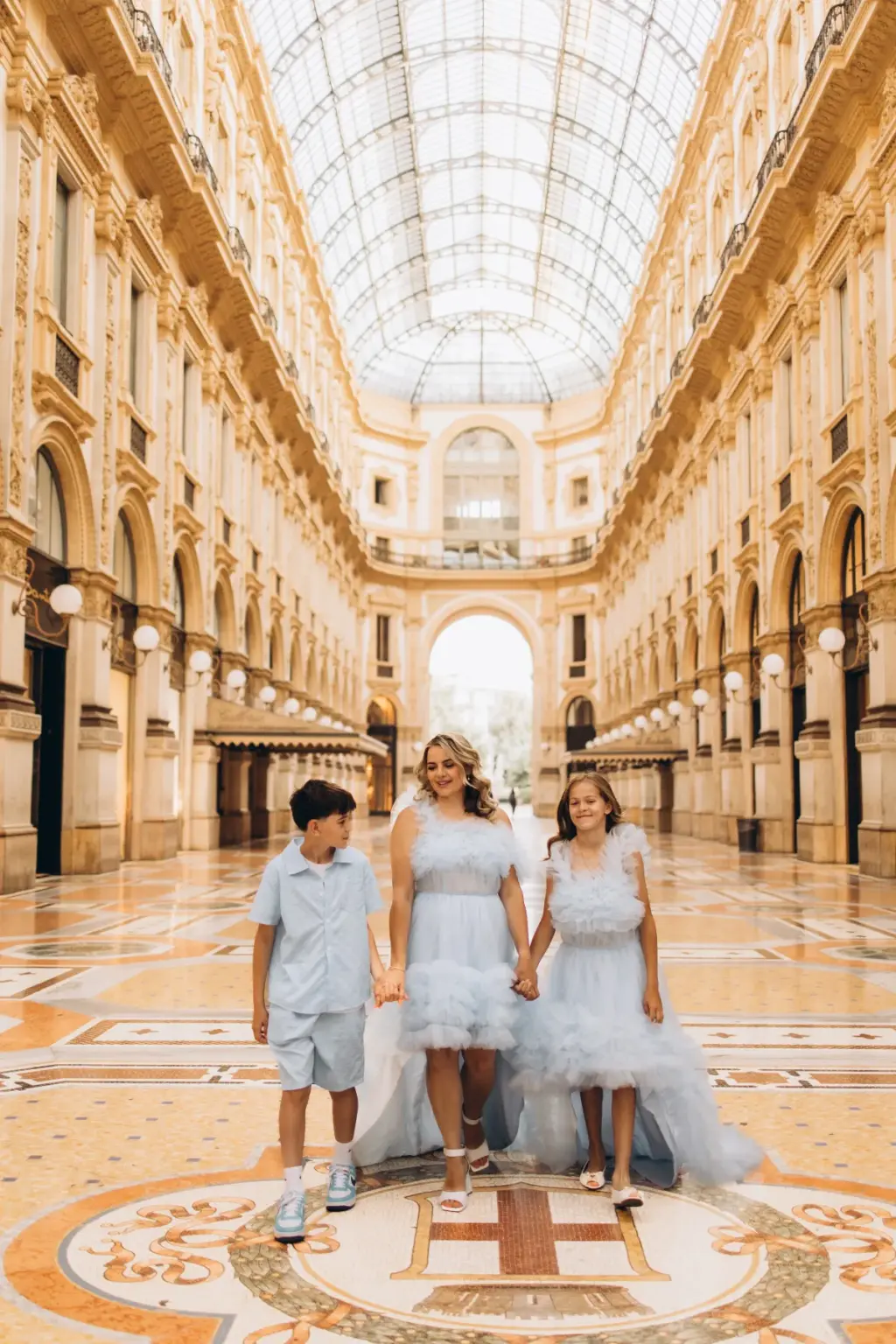 A mother in a pale blue tulle gown walks hand in hand with her son and daughter across the mosaic floor of the Galleria in Milan.