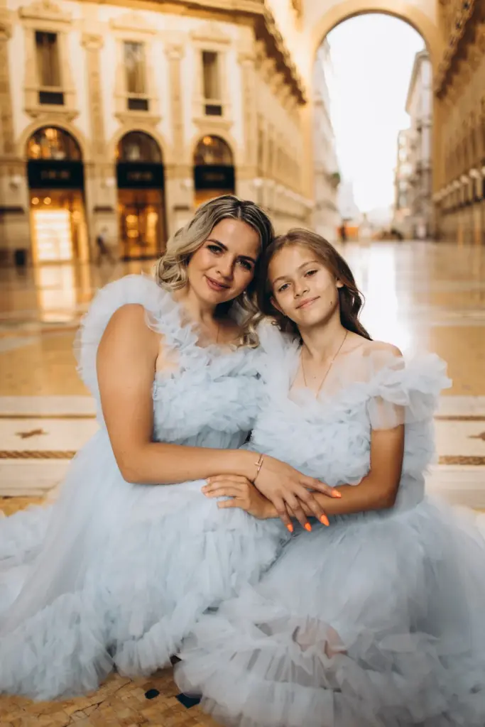 A mother and her daughter in matching pale blue tulle dresses sit cheek to cheek, hands linked, on the floor of the Galleria.