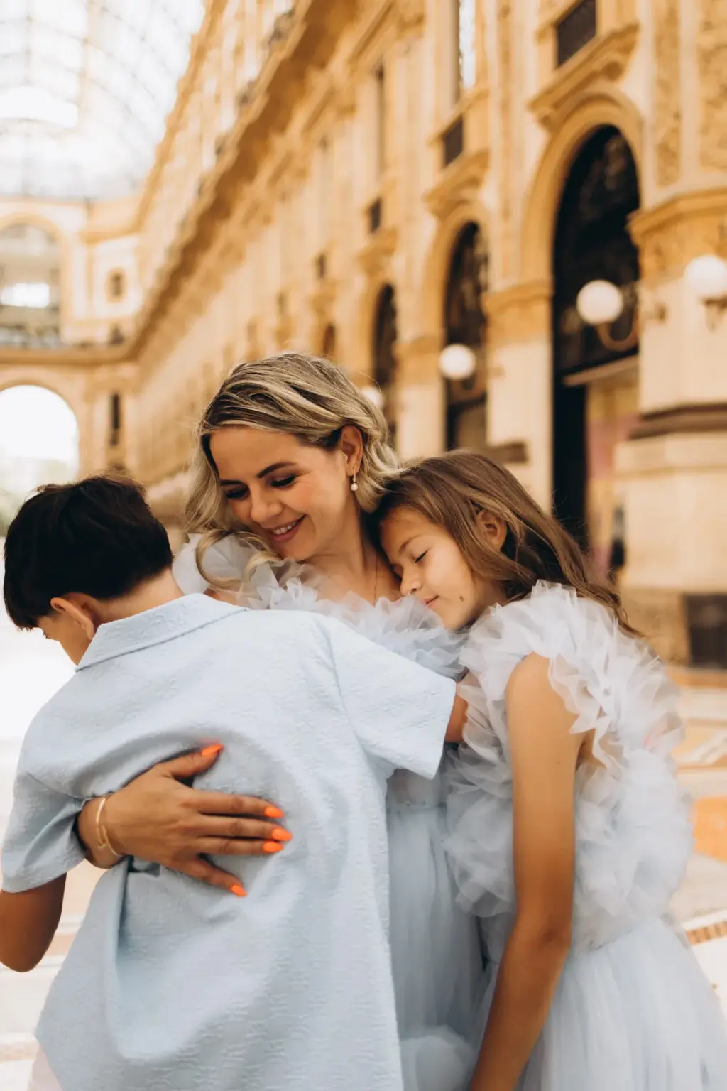 A smiling mother in pale blue tulle hugs her son and daughter close together beneath the arcades of the Galleria in Milan.