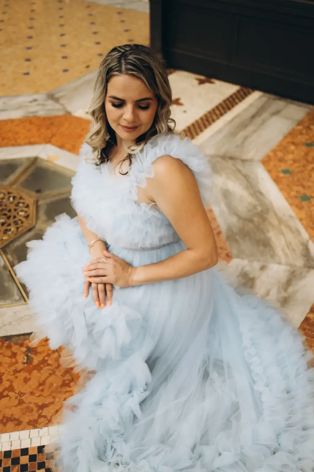 A woman in a ruffled powder blue gown sits on the inlaid marble floor of the Galleria, eyes lowered, hands resting in her lap.