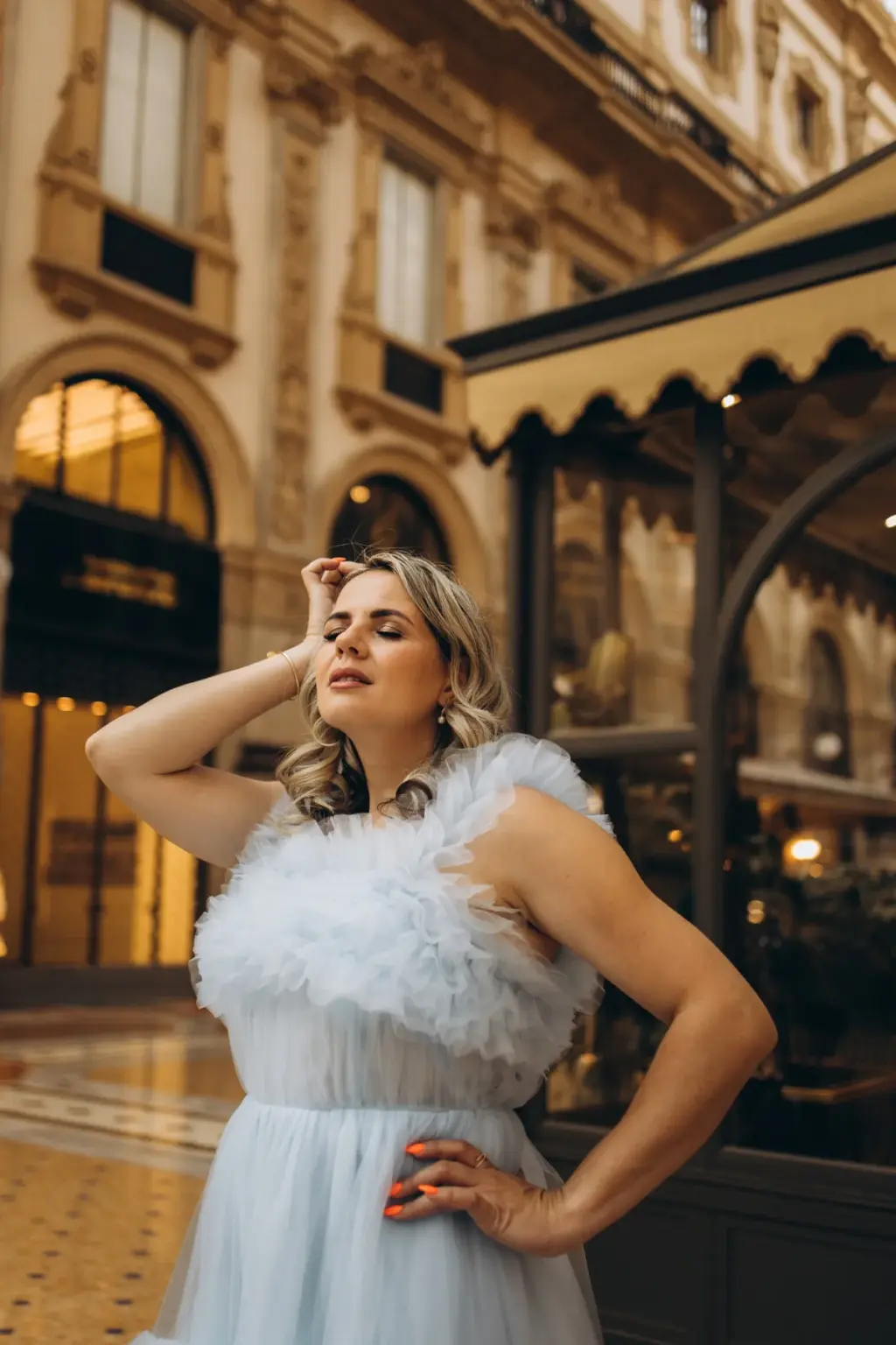 A woman in a pale blue tulle gown tilts her head back with a hand in her hair beside a scalloped cafe awning in the Galleria.