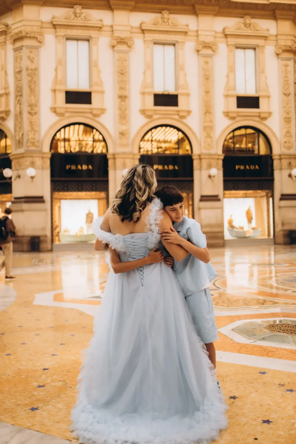 A boy in a light blue shirt hugs his mother from the side as she stands in a pale blue tulle gown on the Galleria's patterned floor.