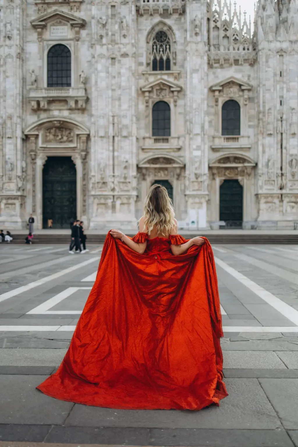 A woman stands with her back to the camera, arms spread to open a red cape-like dress wide before the Duomo's carved doors.