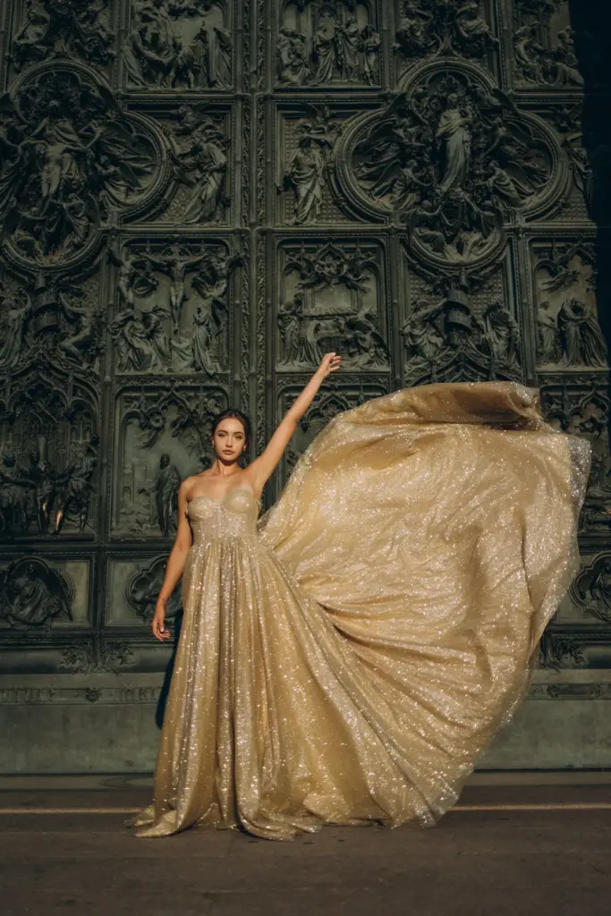 A woman in a gold strapless gown throws one arm up as the fabric flies out sideways against the bronze doors of the Duomo.
