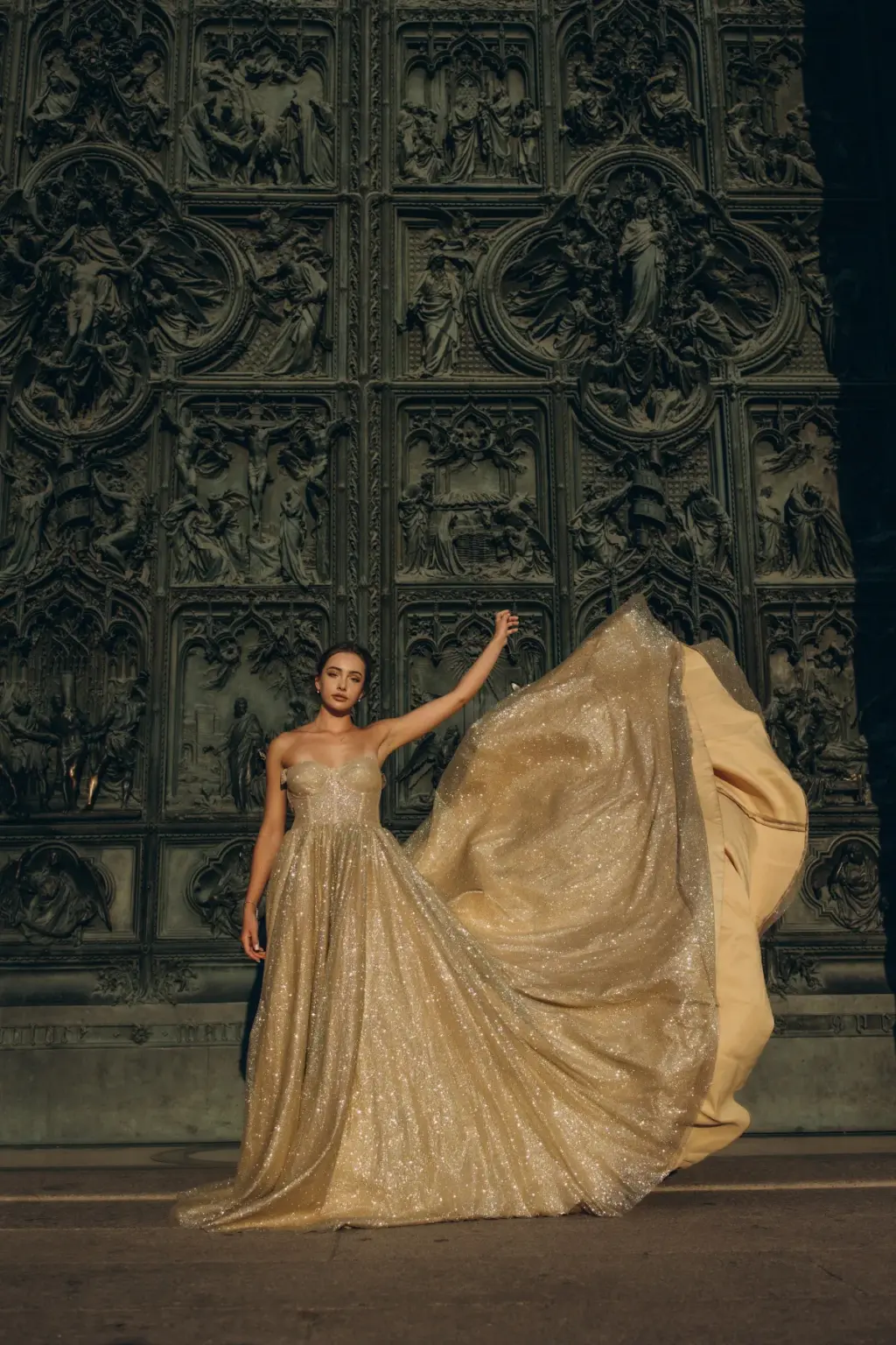 The gold skirt of a flying gown billows into a wide wave as a woman poses before the sculpted bronze panels of the Duomo doors.