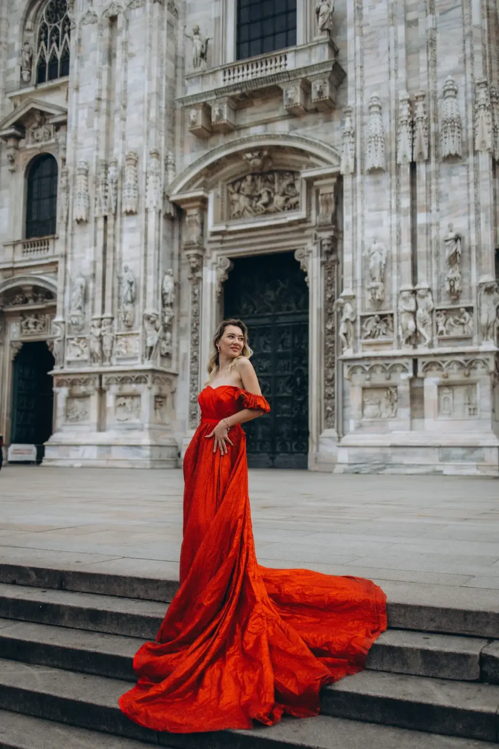 A woman in a strapless red gown smiles from the Duomo steps, her long train draped over the stairs below the bronze doors.