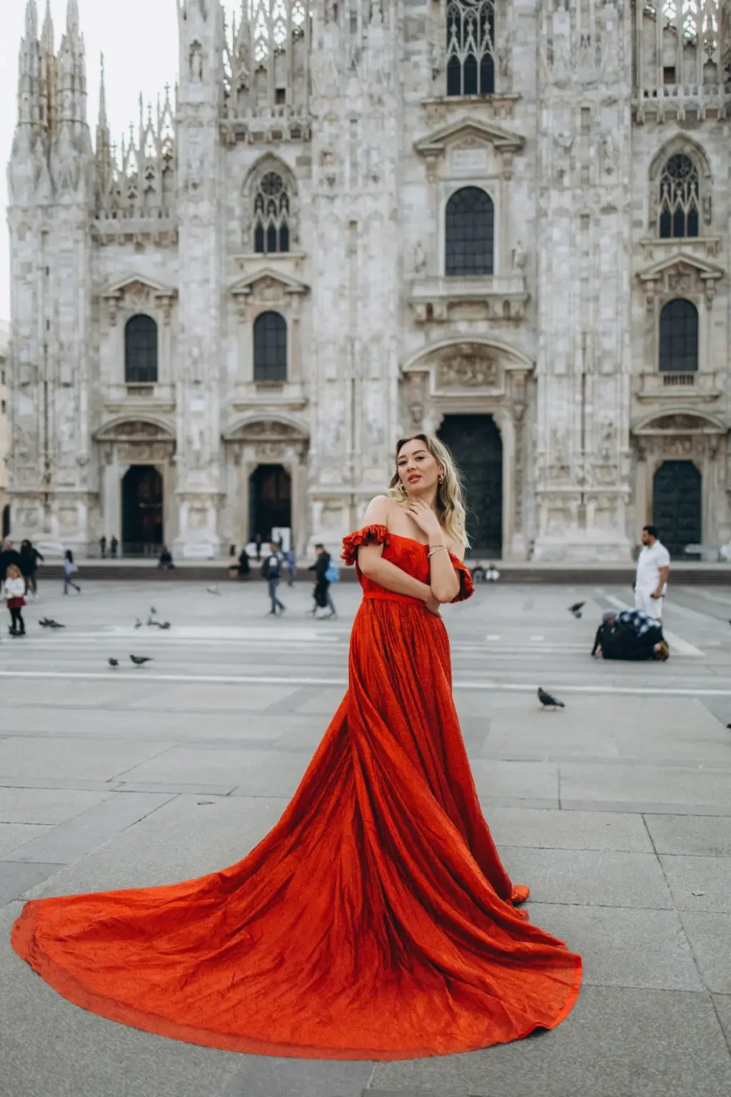 A blonde woman in a long red flying dress looks back over her shoulder, the train pooled on the paving in front of the Duomo di Milano.