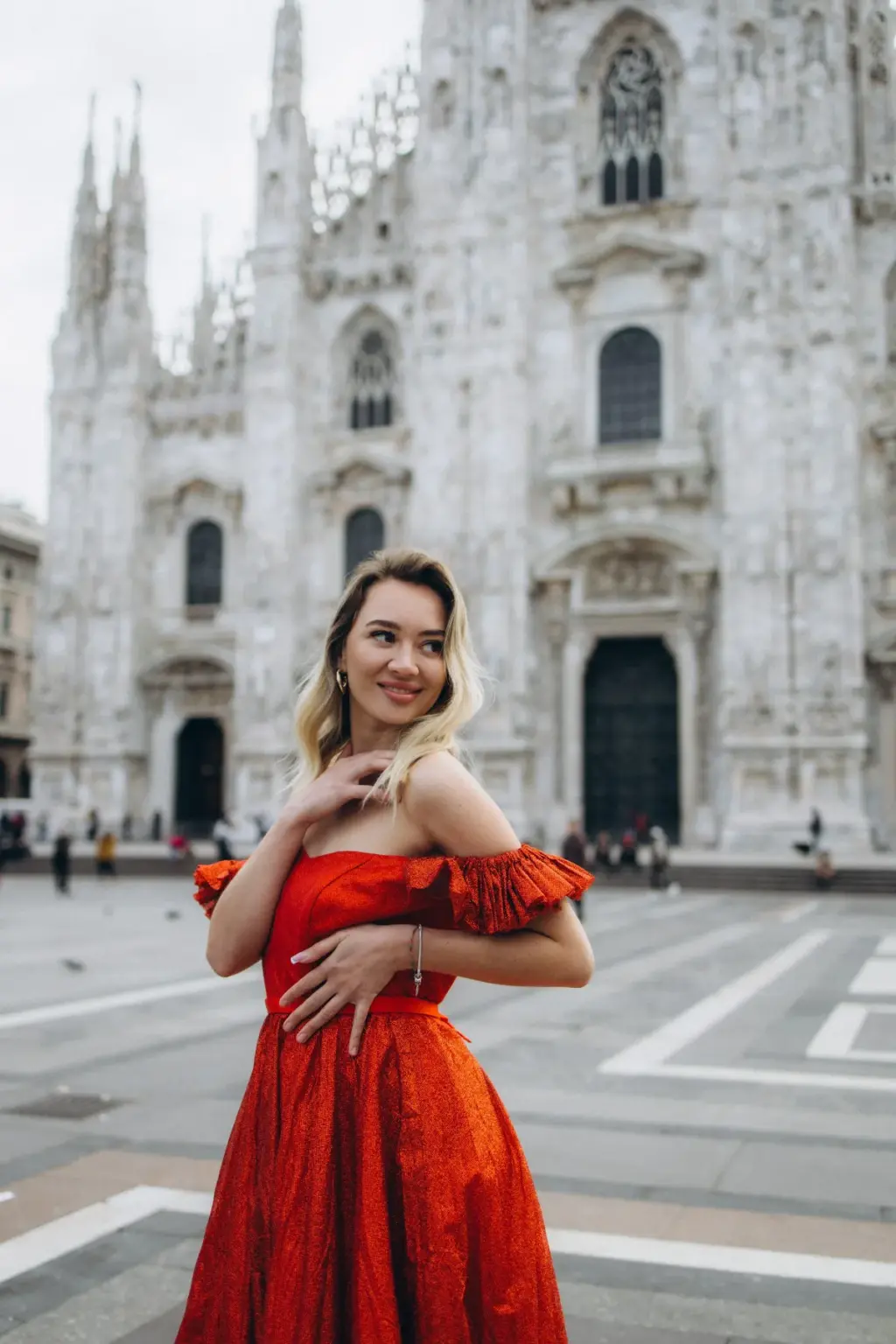 Waist-up portrait of a smiling blonde woman in a ruffled red dress, hand at her collarbone, with the Duomo spires behind her.