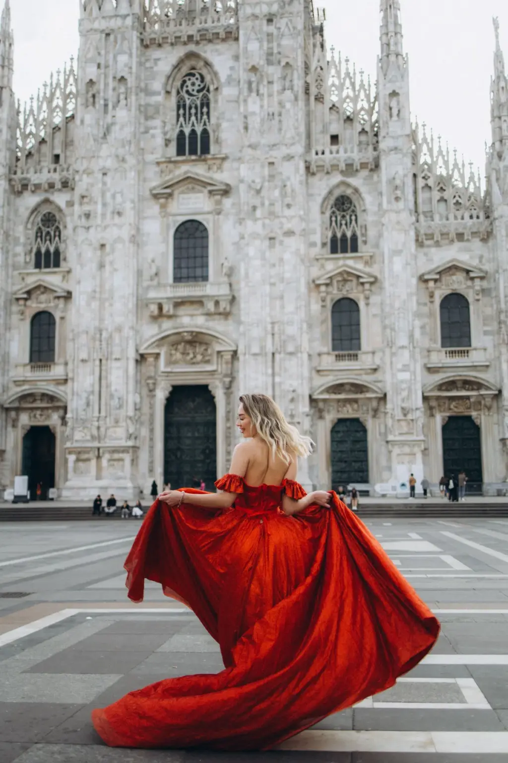 Seen from behind, a woman in a red flying dress swings the skirt out to one side as she faces the facade of the Duomo.