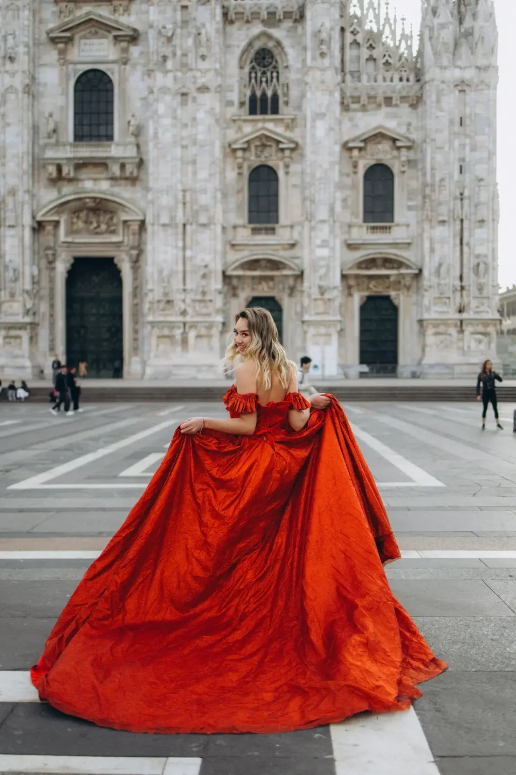 A smiling woman glances back while holding the wide red skirt of her gown open across the striped paving of the Duomo square.