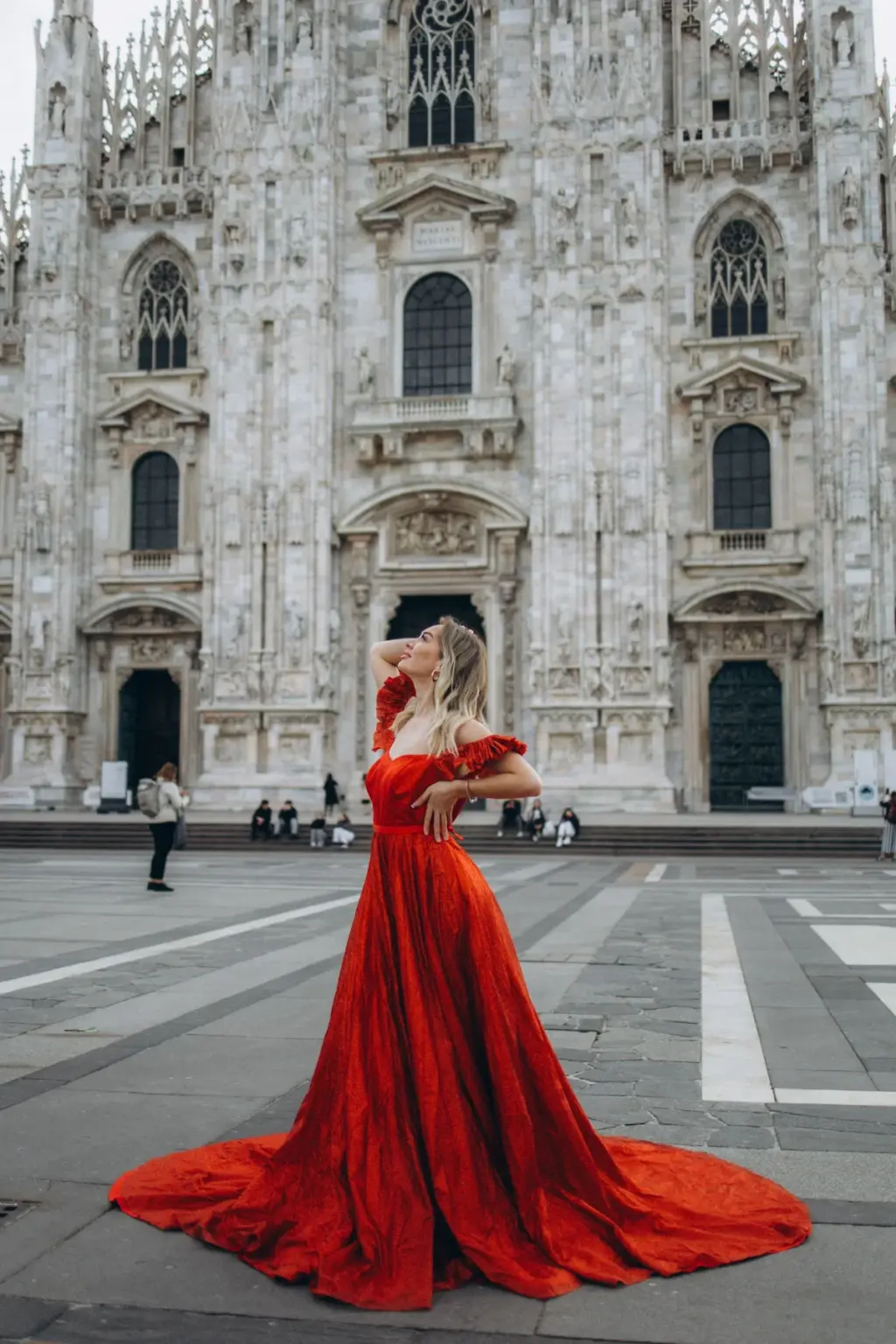 A woman in an off-the-shoulder red gown lifts a hand to her hair as the long train spreads around her on Piazza del Duomo.