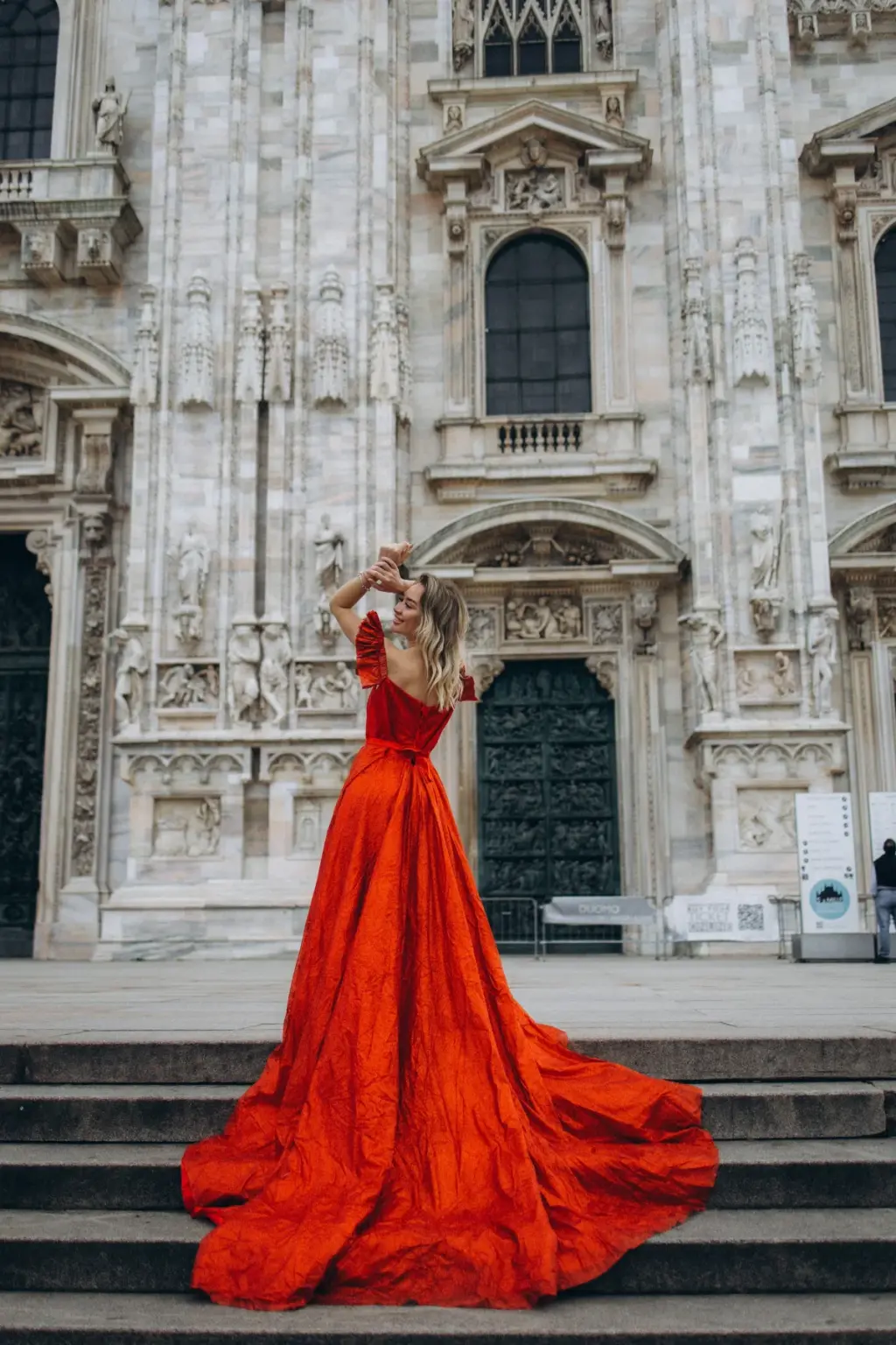 A woman in a red gown stands on the Duomo steps with both hands in her hair, the train cascading down the stone stairs.