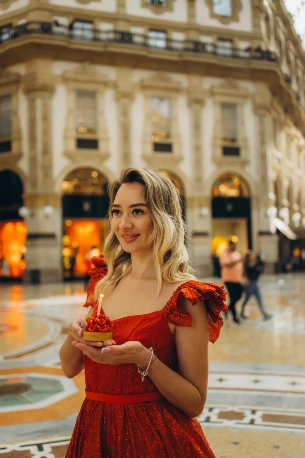 A woman in a red sequin dress holds a small red cake with a lit candle in her palms inside the Galleria Vittorio Emanuele II.