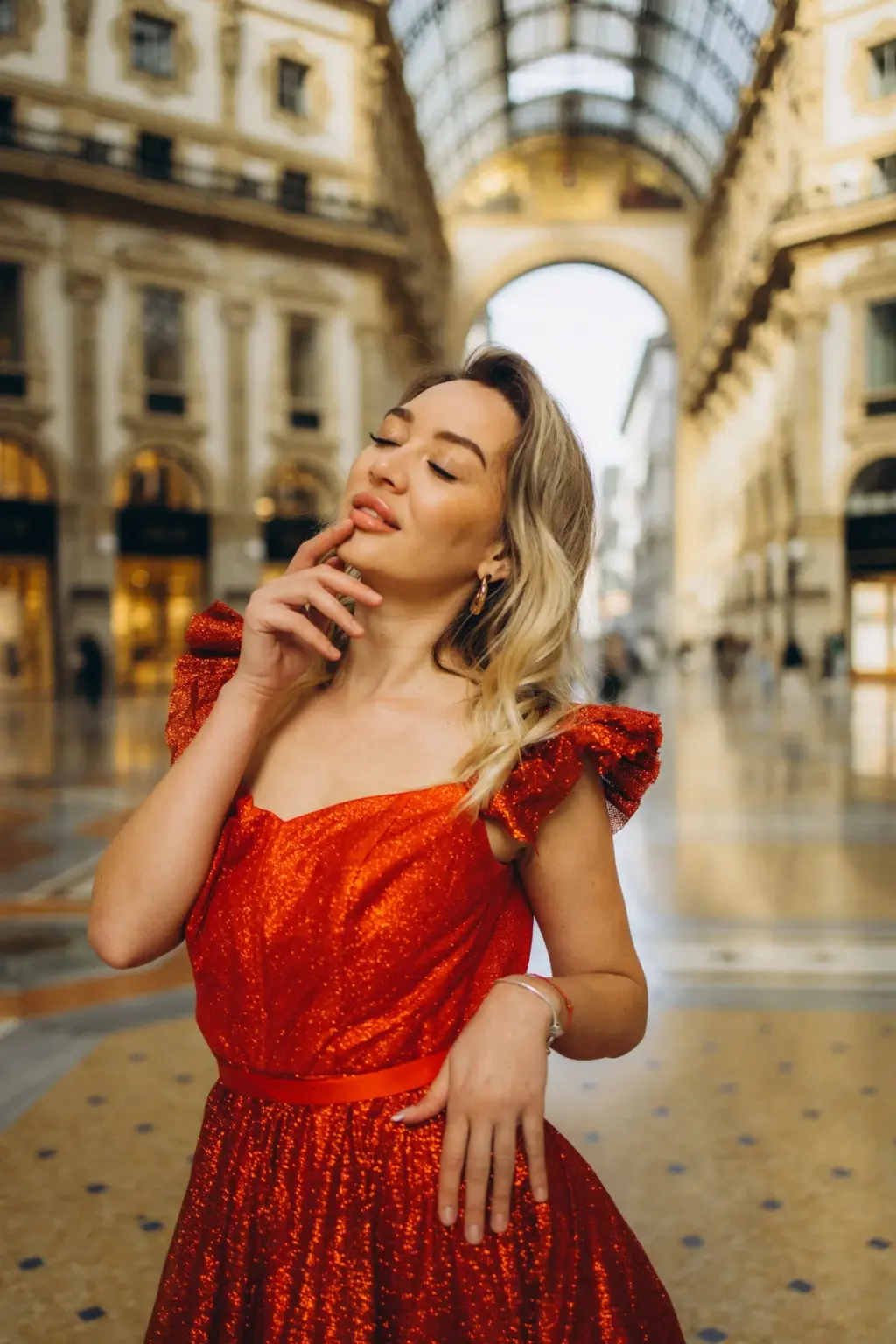 Close portrait of a woman with closed eyes and a finger at her lips in a red ruffled dress, the Galleria arch blurred behind her.