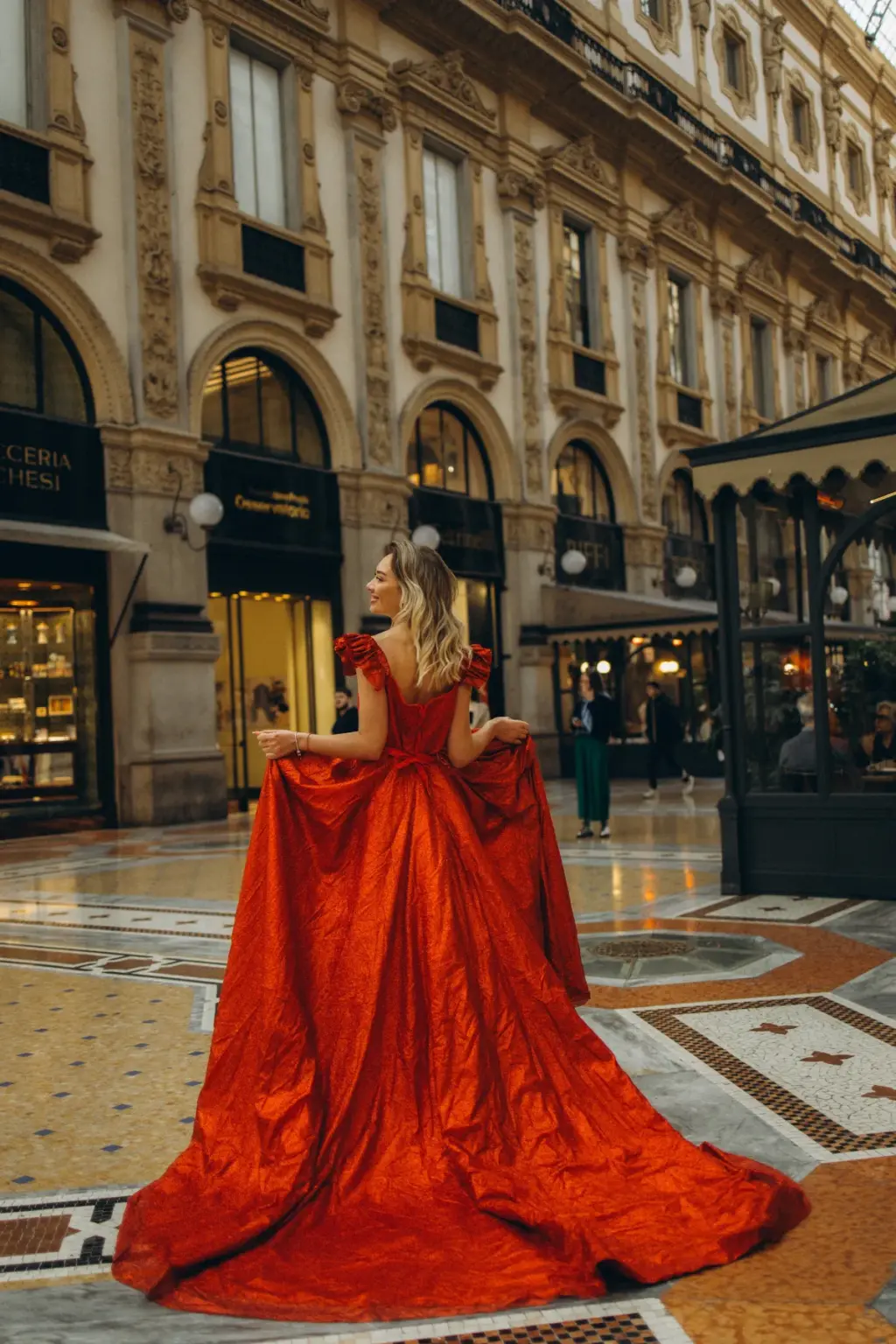 From behind, a woman holds out both sides of her red gown as the long skirt sweeps over the Galleria mosaic past the shop arcades.