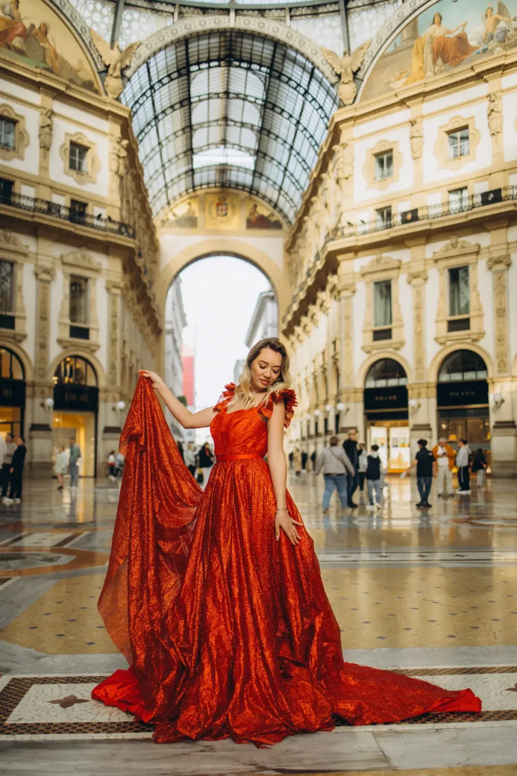 A woman lifts one side of her red sequin gown high beneath the glass dome and arched entrance of the Galleria Vittorio Emanuele II.