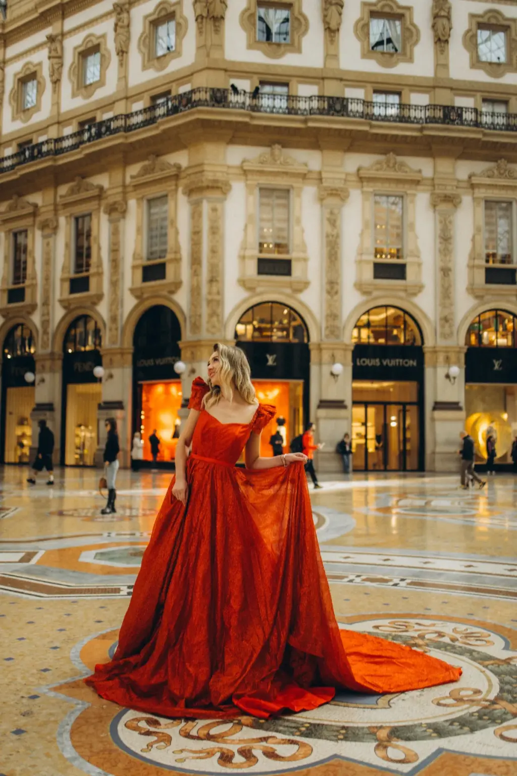 A woman in a sparkling red gown stands on the mosaic floor at the centre of the Galleria, holding the skirt as it fans out around her.