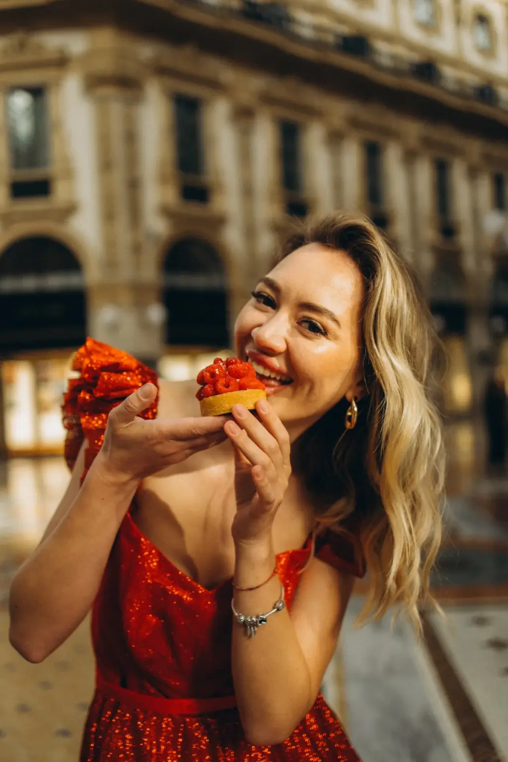 A laughing woman in a red sequin dress takes a bite of a small red pastry, with the arcades of the Galleria softly blurred behind.