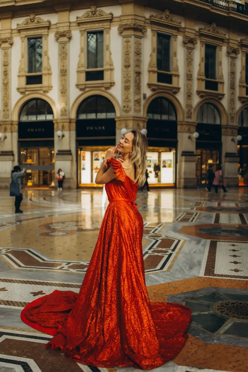 A woman in a glittering red gown turns her shoulder to the camera, the train spread over the patterned marble of the Galleria floor.