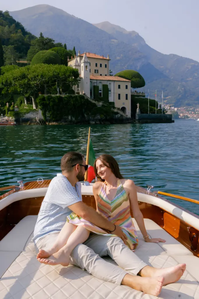 A couple on a wooden boat with an Italian flag, Villa del Balbianello behind them