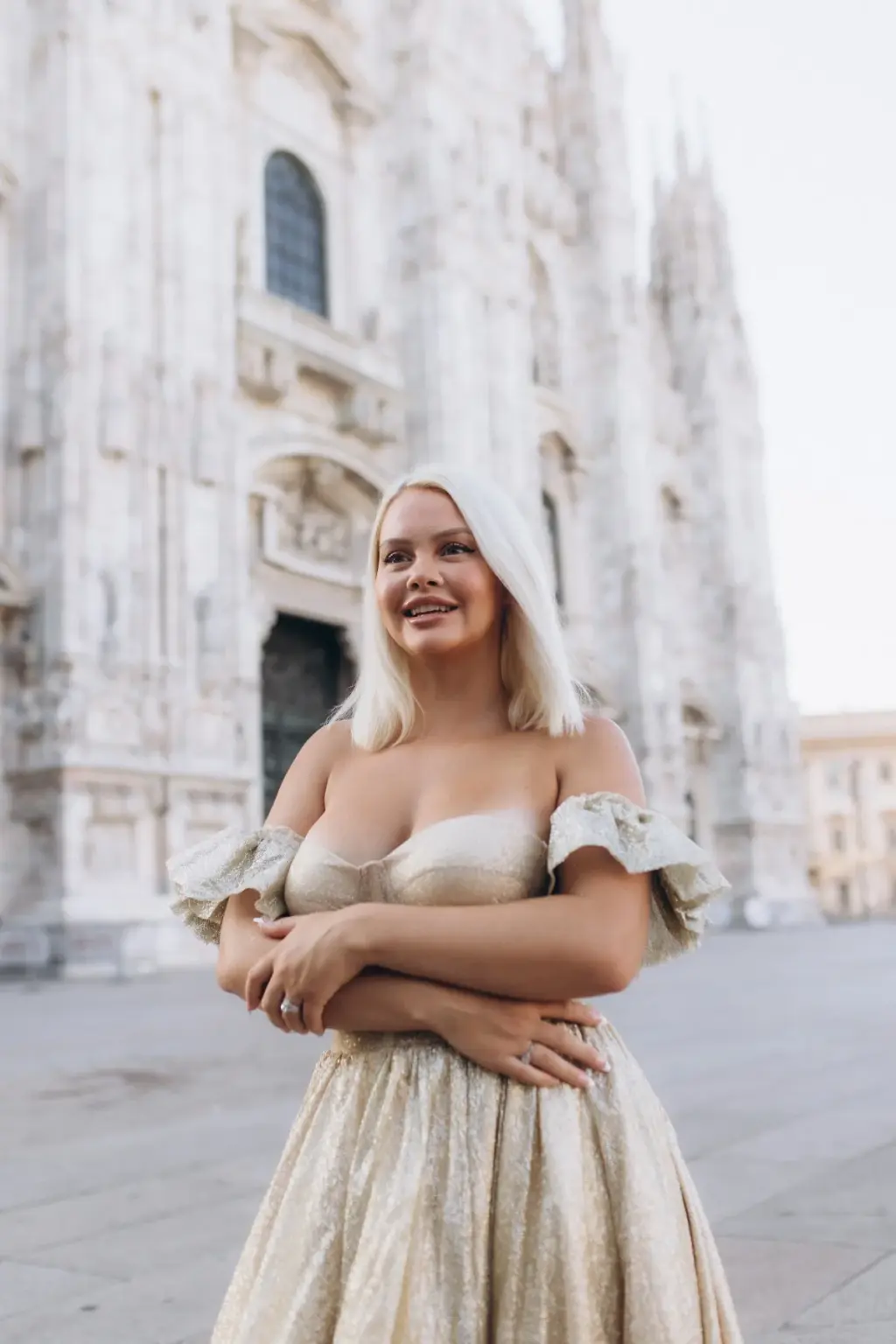 A smiling platinum-blonde woman hugs her arms around her gold gown, looking off to the side with the Duomo facade behind her.