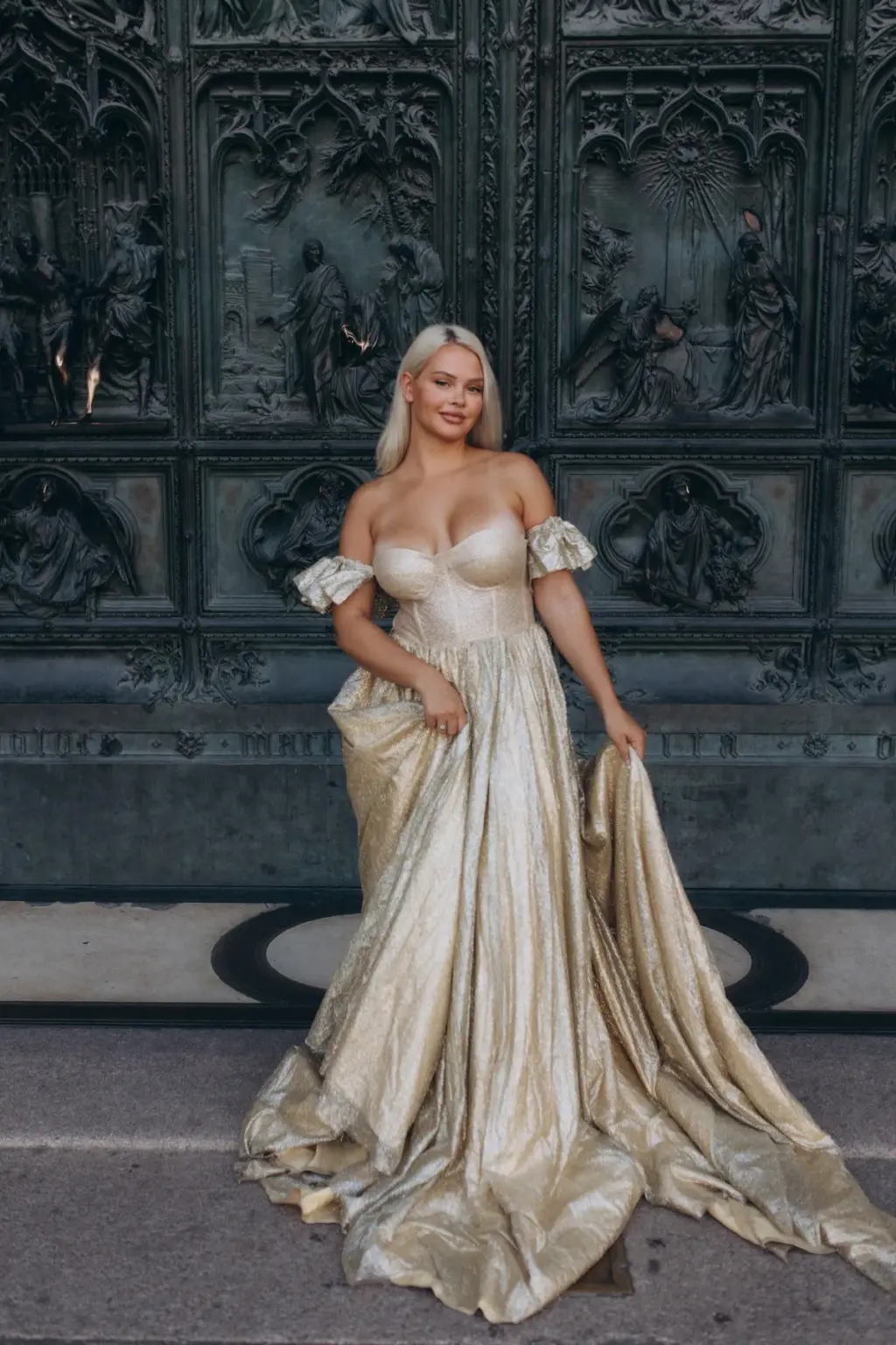 A blonde woman in a strapless gold gown holds out her skirt in front of the sculpted bronze doors of the Duomo di Milano.
