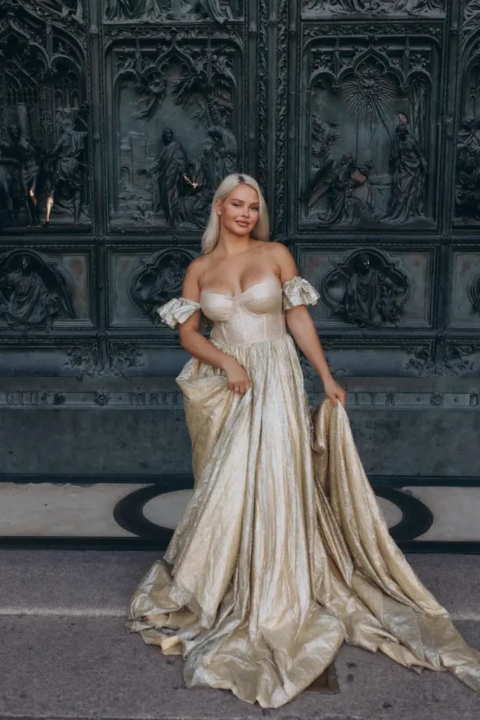 A blonde woman in a strapless gold gown holds out her skirt in front of the sculpted bronze doors of the Duomo di Milano.
