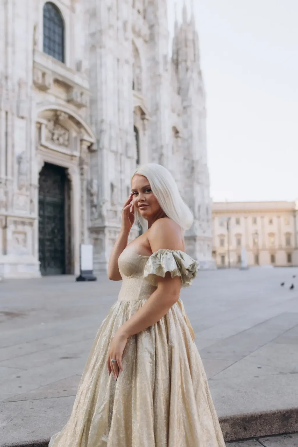A woman with platinum hair in a champagne off-the-shoulder gown touches her cheek on the quiet square beside the Duomo di Milano.