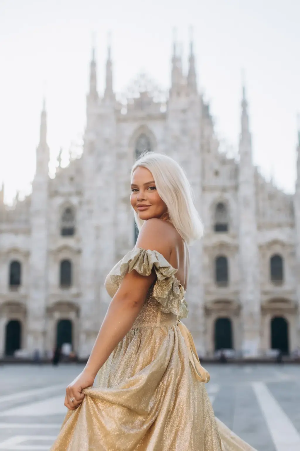 A platinum-blonde woman twirls her gold gown and smiles back at the camera on the square in front of the Duomo's spires.