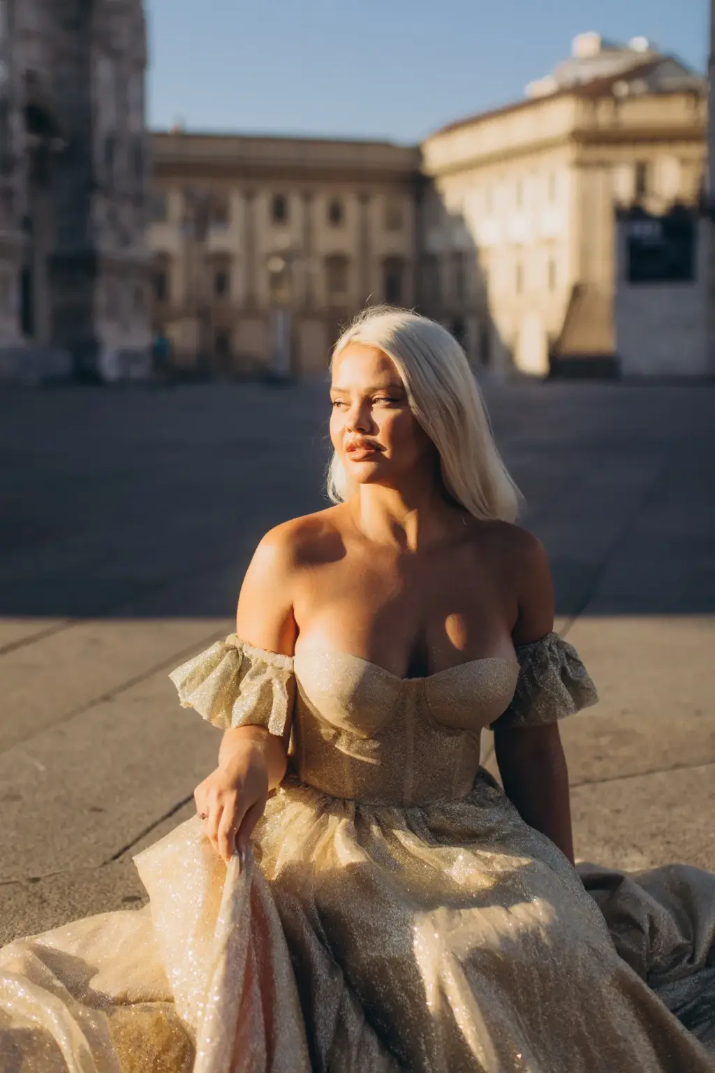 A platinum-blonde woman sits on the paving in a gold gown, face turned toward the sun, with the square's buildings behind.